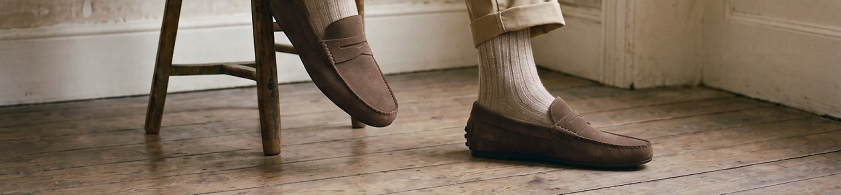 A person wearing men's brown European leather suede penny loafers, cream ribbed socks, and khaki trousers, sitting on a wooden stool in a room with a rustic wood floor and white wall.