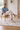 A man sits in a wooden chair on a white patterned rug in a bright, modern living room. He is wearing a light blue button-down shirt, cream trousers, and men's beige Italian suede leather sneaker dress shoes.