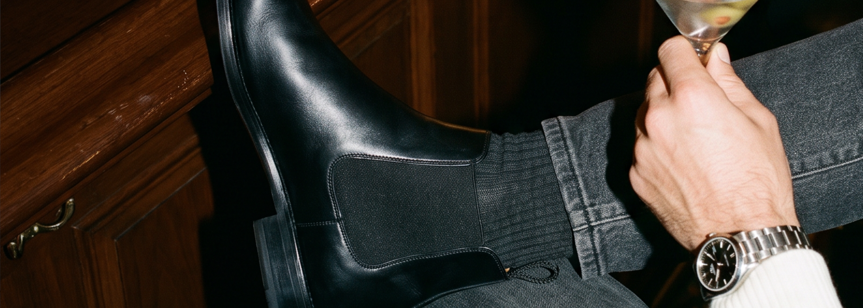 A close-up lifestyle shot of a man wearing a men's black Italian leather Chelsea boot, dark gray jeans, and a silver watch, holding a cocktail glass in a dimly lit room with dark wood paneling.