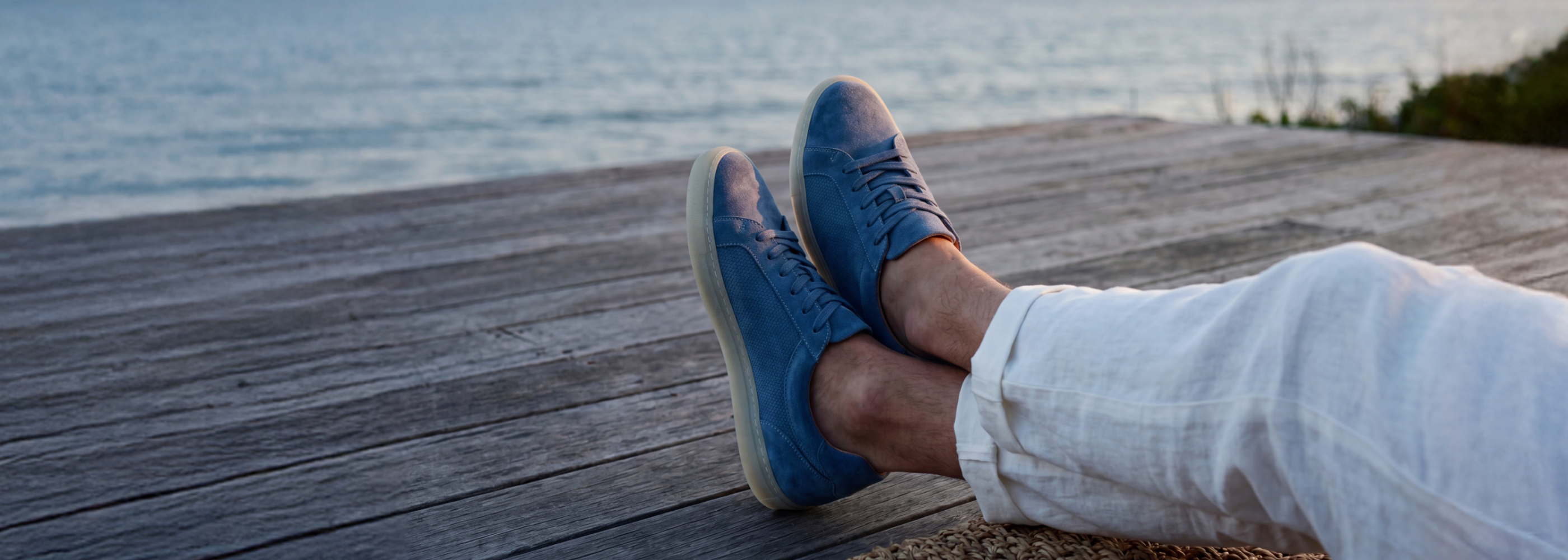 Feet wearing men's european blue suede leather dress sneakers and white pants on a wooden deck by the water.